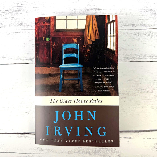 Book cover of 'The Cider House Rules' by John Irving with a blue chair in a room.