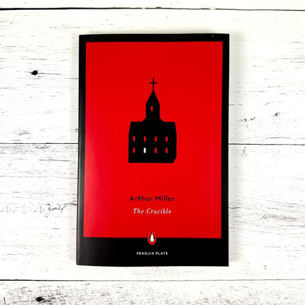 paperback copy of 'The Crucible' by Arthur Miller with red book cover and silhouette of a church on a white wood surface