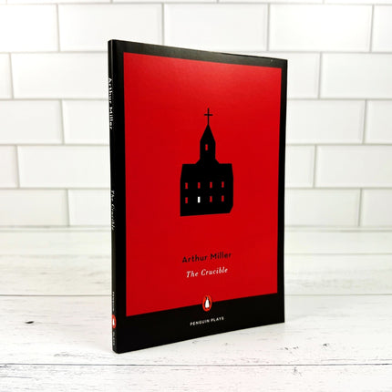 Book titled 'The Crucible' by Arthur Miller with a red cover and black silhouette of a building on a white tiled background.