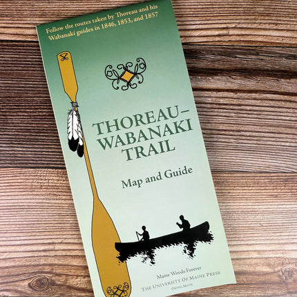 a folded green map against a wood background with a large canoe paddle adorned with feathers next to two silhoutted figures paddling a canoe with the title "Thoreau-Wabanaki Trail - Map and Guide"