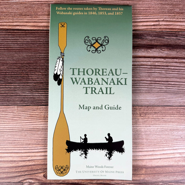 a folded green map against a wood background with a large canoe paddle adorned with feathers next to two silhoutted figures paddling a canoe with the title "Thoreau-Wabanaki Trail - Map and Guide"