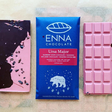 Three Ursa Major bars side by side. The center bar is in its royal blue wrapper with ursa major constellation illustration and the two surrounding bars are unwrapped and showing off their brilliant pink color swirled with dark chocolate and speckled with cacao nibs