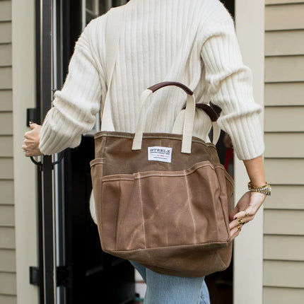Person holding a brown tote bag with a visible brand label, wearing a white sweater and light blue jeans.