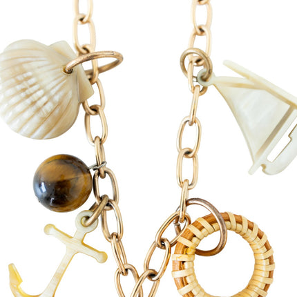 Gold charm necklace with various charms including a shell, anchor, and tiger's eye stone on a white background.