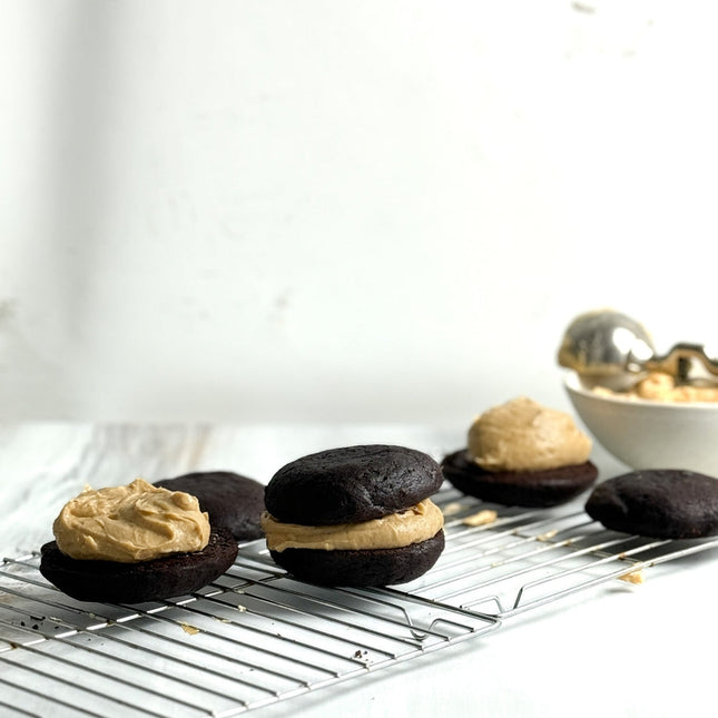 Chocolate cake or cookie whooperas filled with peanut butter cream, arranged on a metal cooling rack with a bowl of more cream in the background.