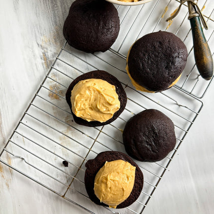 Four chocolate cupcakes, including two topped with swirls of creamy peanut butter frosting, arranged on a wire cooling rack over a white wooden surface. A bowl of peanut butter frosting and a metal spatula are visible in the background.