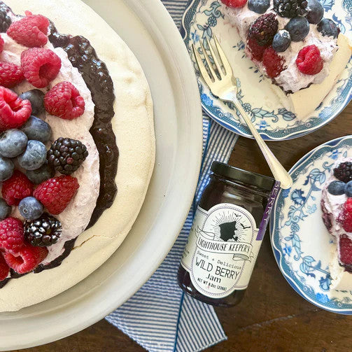 a jar of wild berry jam laying on a table alongside a frosted cake on a platter that is piled high with fresh berries and a couple plates of sliced cake