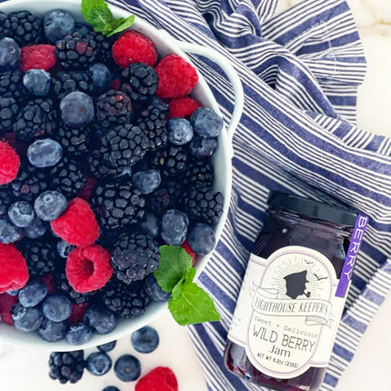 a jar of wild berry jam next to a bowl overflowing with fresh blackberries, raspberries, blueberries laid out on a blue and white striped cloth