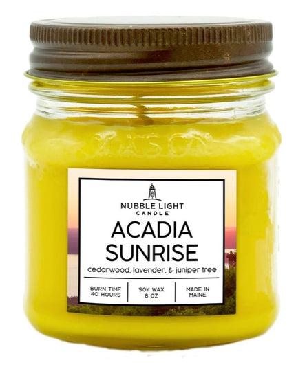 a candle in a clear glass mason jar with bright yellow wax and a label that reads "Acadia Sunrise" against a white background