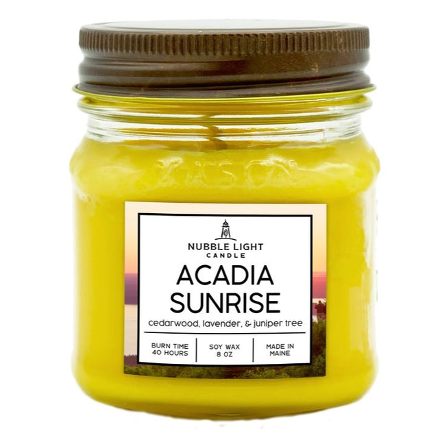 a candle in a clear glass mason jar with bright yellow wax and a label that reads "Acadia Sunrise" against a white background