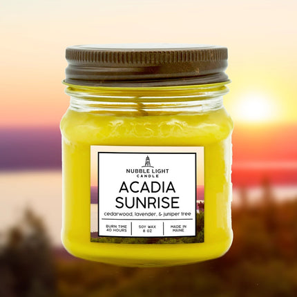 a candle in a clear glass mason jar with bright yellow wax and a label that reads "Acadia Sunrise" with an image of an ocean sunrise behind it