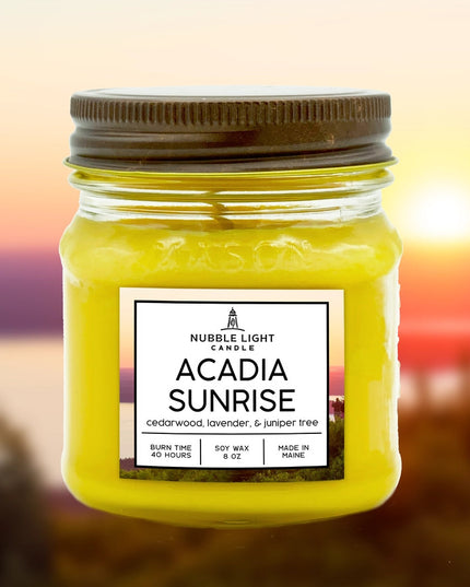 a candle in a clear glass mason jar with bright yellow wax and a label that reads "Acadia Sunrise" with an image of an ocean sunrise behind it