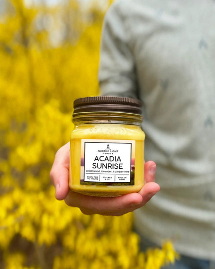 the acadia sunrise candle held in a model's hand with yellow blooming hedges in the background