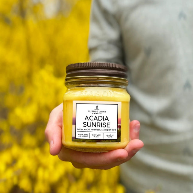 the acadia sunrise candle held in a model's hand with yellow blooming hedges in the background