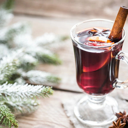 a wintry themed tabletop with balsam boughs and an elegant clear glass mug containing a cranberry-red  mulled beverage, garnished with a citrus, spices, and cinnamon stick
