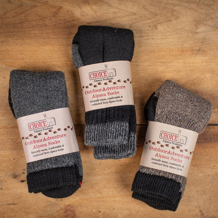 three pairs of alpaca outdoor adventure socks folded in their packaging on a wood background showing the available colors for the socks - gray, black, and cocoa brown
