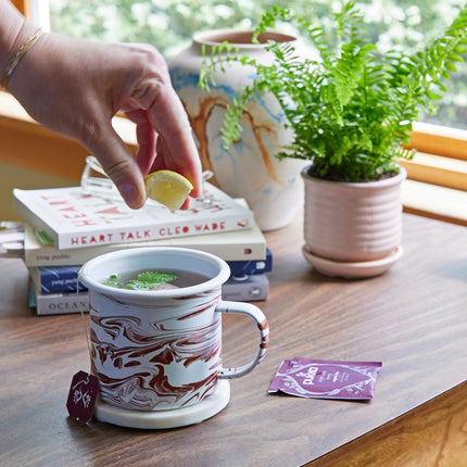The mostly medicinal lemon balm altered herbs mug on a tabletop next to a potted fern. the mug is filled with steaming tea and some lemon balm leaves with a tea bag package nearby - a hand hovers over the mug squeezing a fresh lemon slice into the tea