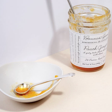 A nearly empty jar of Bonnie's Peach Ginger jam next to a white bowl with a silver spoon coated in jam