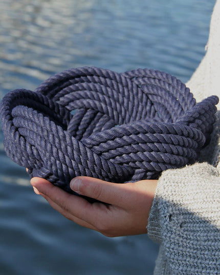 a hand woven cotton rope bowl in navy blue seen in profile in the hands of someone standing at the waterfront