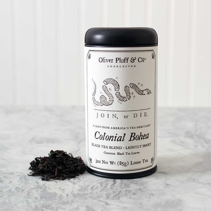 Oliver Pluff & Co Colonial Bohea tea tin with Join or Die political cartoon by Benjamin Franklin on the label. Tea Tin stands on a white marble surface with a heap of loose black tea leaves next to it against a white background