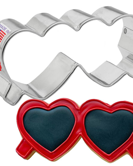 a silver, tin cookie cutter in the shape of folded heart-shaped glasses next to a fully decorated cookie of folded heart shaped sunglasses. the frames are decorated with red icing and the lenses of the glasses have black icing.