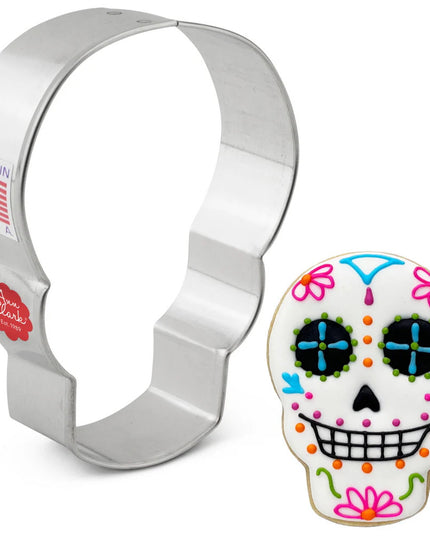 silver tin cookie cutter in the shape of a skull next to a fully decorated cookie of a skull with a toothy smile and frosted with white icing and colorful, intricate details following the aesthetic traditions of day of the dead sugar skull decor