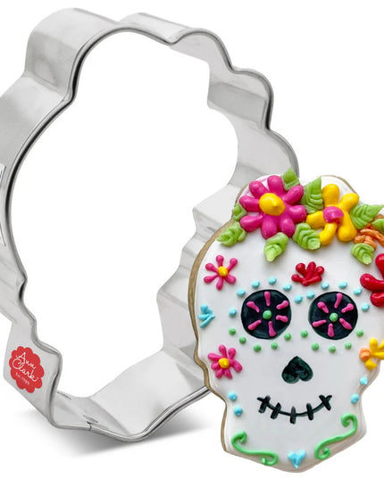 silver tin cookie cutter in the shape of a skull with flowers on top of the head next to a fully decorated cookie of a skull with flowers on top of its head with white icing and colorful, intricate details following the aesthetic traditions of day of the dead sugar skull decor and a black top hat with intricate, colorful decor