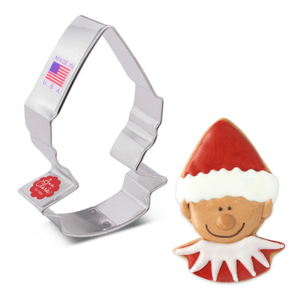 A tin cookie cutter in the shape of an elf's head with a pointed elf hat next to a fully decorated cookie in the shape of an elf's head with a pointed hat iced with red icing and white trim on the hat, a smiling elf face with medium skin tone, and a white pointed collar on a red shirt