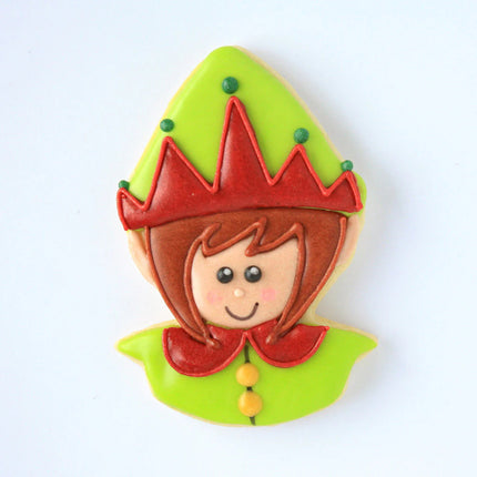 a fully decorated cookie in the shape of an elf's head with a pointed hat iced with green icing and red decorative detail on the hat, a smiling elf face with fair skin tone, and a red, rounded collar on a green shirt