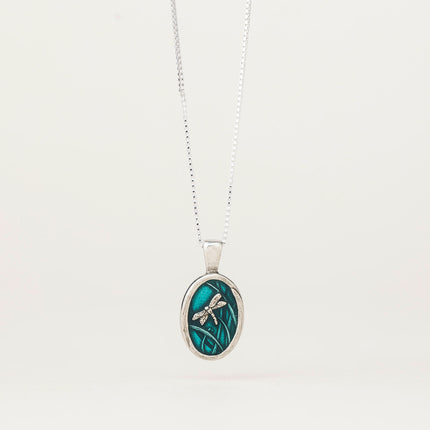 an oval pewter pendant with a teal oval inner with an engraved dragonfly and reeds hanging from a silver chain