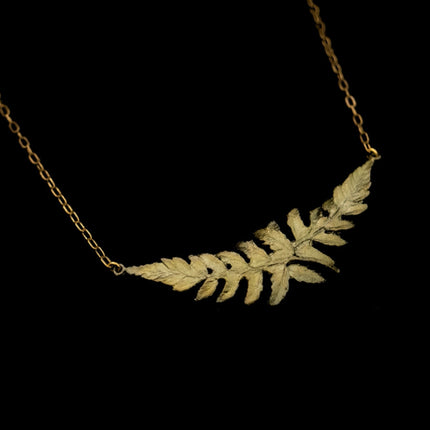 an angled close up view of a delicate chain with a pendant featuring two overlapping fern sprigs cast in bronze with a delicate green patina