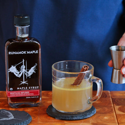 a bar scene with a drink being mixed - a bottle of festivus infused maple syrup sits on the bar next to a rocks glass filled with a cider like beverage and a cinnamon stick, showing that the syrup has versatile use for cocktails
