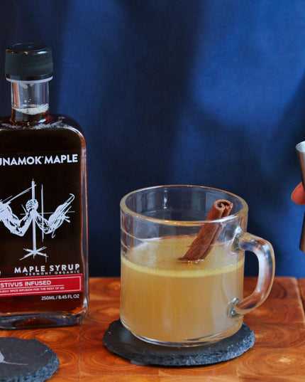 a bar scene with a drink being mixed - a bottle of festivus infused maple syrup sits on the bar next to a rocks glass filled with a cider like beverage and a cinnamon stick, showing that the syrup has versatile use for cocktails