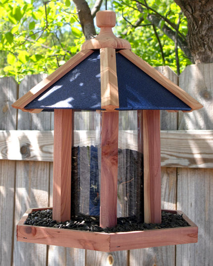 a backyard scene with the gazebo feeder hanging from a tree with a wood fence behind it. the feeder is hexagonal in shape with a blue roof