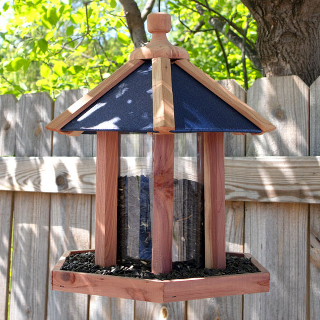 a backyard scene with the gazebo feeder hanging from a tree with a wood fence behind it. the feeder is hexagonal in shape with a blue roof