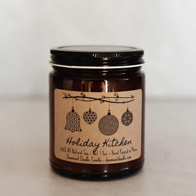 a candle in an amber glass jar with black lid and kraft label with handwriting style graphics that read "Holiday Kitchen" below various shapes of hand drawn holiday ornaments