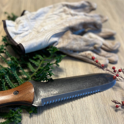 Essential Hori Hori knife unsheathed on a surface alongside some freshly cut greens with gardening gloves