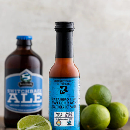 a bottle of habanero lime switchback craft beer hot sauce on a dark wood table surrounded by whole and sliced limes with a bottle of switchback ale in the background