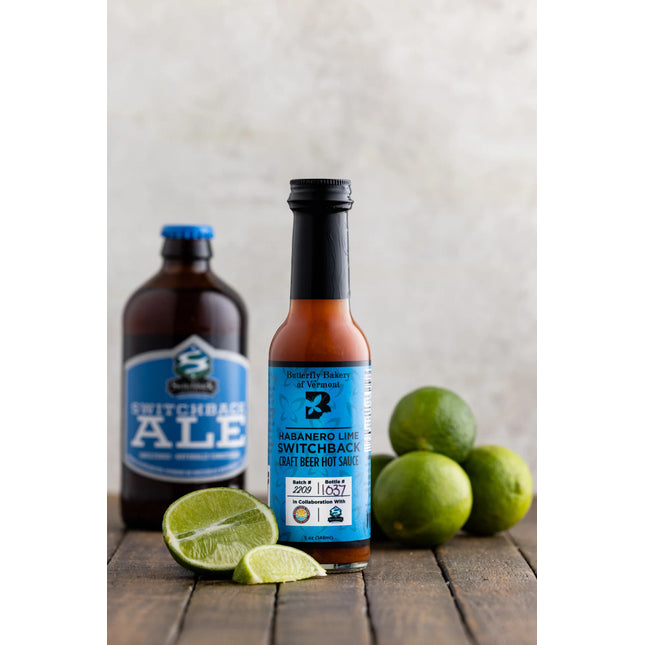 a bottle of habanero lime switchback craft beer hot sauce on a dark wood table surrounded by whole and sliced limes with a bottle of switchback ale in the background