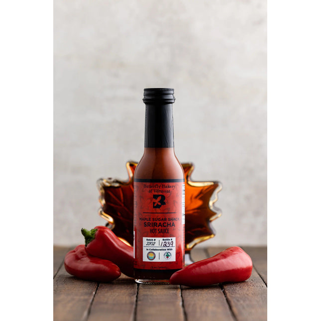 a bottle of maple sugar shack sriracha on a dark wood table surrounded by red serrano and jalepeno peppers with a maple leaf shaped bottle of maple syrup behind it