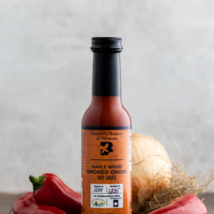 a bottle of smoked onion hot sauce on a dark wood table surrounded by red serrano and jalapeño peppers with a large onion behind it