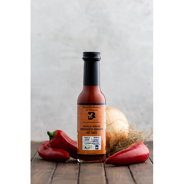 a bottle of smoked onion hot sauce on a dark wood table surrounded by red serrano and jalapeño peppers with a large onion behind it