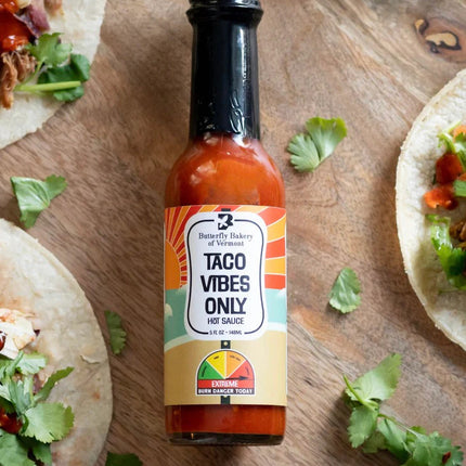a bottle of taco vibes only hot sauce laid on a wood table surrounded by a sprinkling of cilantro leaves with the edges of tacos visible near the borders of the image - the bottle has a heat meter on it noting that this sauce has "Extreme Heat"