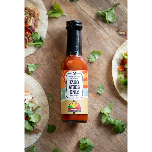 a bottle of taco vibes only hot sauce laid on a wood table surrounded by a sprinkling of cilantro leaves with the edges of tacos visible near the borders of the image - the bottle has a heat meter on it noting that this sauce has "Extreme Heat"