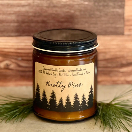 a candle in an amber glass jar with black lid and kraft label with handwriting style graphics that read "Knotty Pine" above a silhouetted forest of pine trees flanked by evergreen sprigs