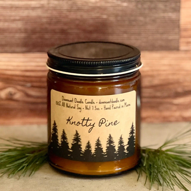 a candle in an amber glass jar with black lid and kraft label with handwriting style graphics that read "Knotty Pine" above a silhouetted forest of pine trees flanked by evergreen sprigs