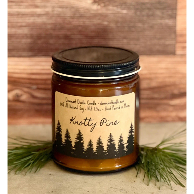 a candle in an amber glass jar with black lid and kraft label with handwriting style graphics that read "Knotty Pine" above a silhouetted forest of pine trees flanked by evergreen sprigs