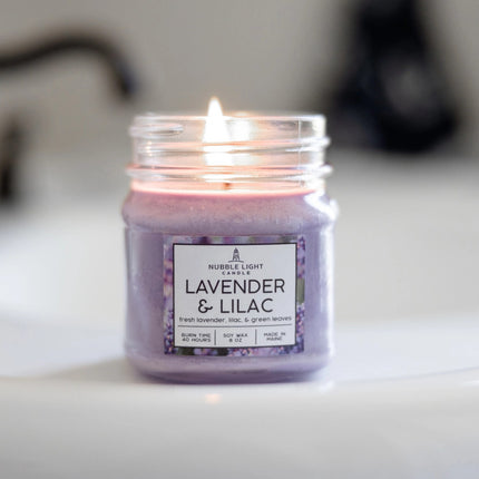 a candle in a clear glass mason jar with light purple wax and a label that reads "Lilac & Lavender" on a bathroom sink