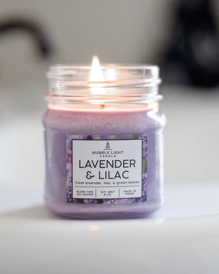 a candle in a clear glass mason jar with light purple wax and a label that reads "Lilac & Lavender" on a bathroom sink