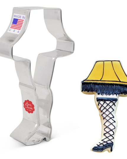 a tin cookie cutter in the shape of a leg in high heels topped with a lampshade next to a fully decorated cookie in the same shape and frosted with yellow icing on the lampshade with black fringe, a black high heeled shoe on the leg, and fishnet stockings on the leg